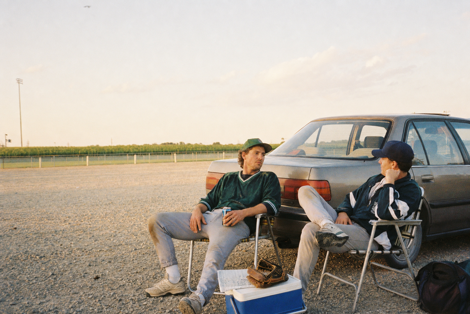 Two friends in vintage sportswear reclining in lawn chairs behind a sedan at golden hour