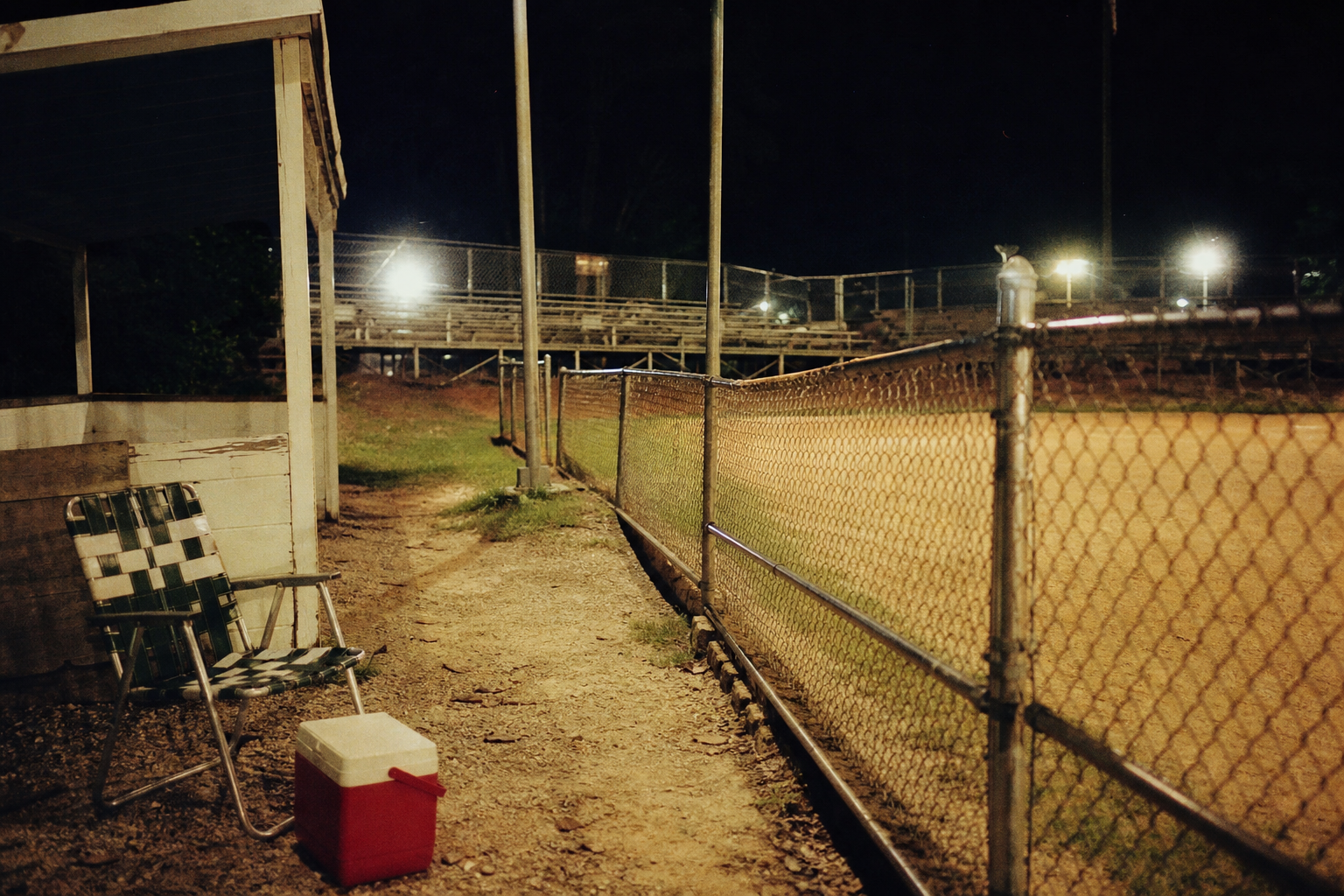Empty lawn chair and cooler beside a chain-link fence at a small-town ballpark at night